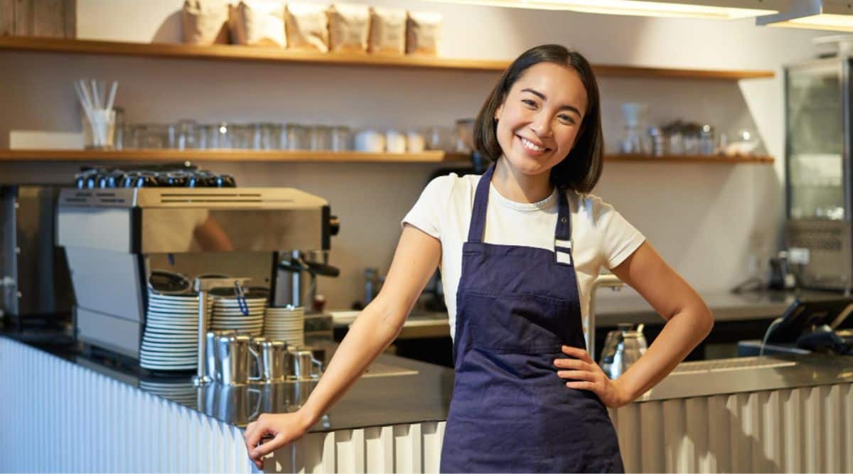 A smiling young barista wearing an apron works in a café, standing near the counter with a coffee machine, while also considering the importance of "Market Research for Small Businesses" to ensure customer satisfaction and business success.